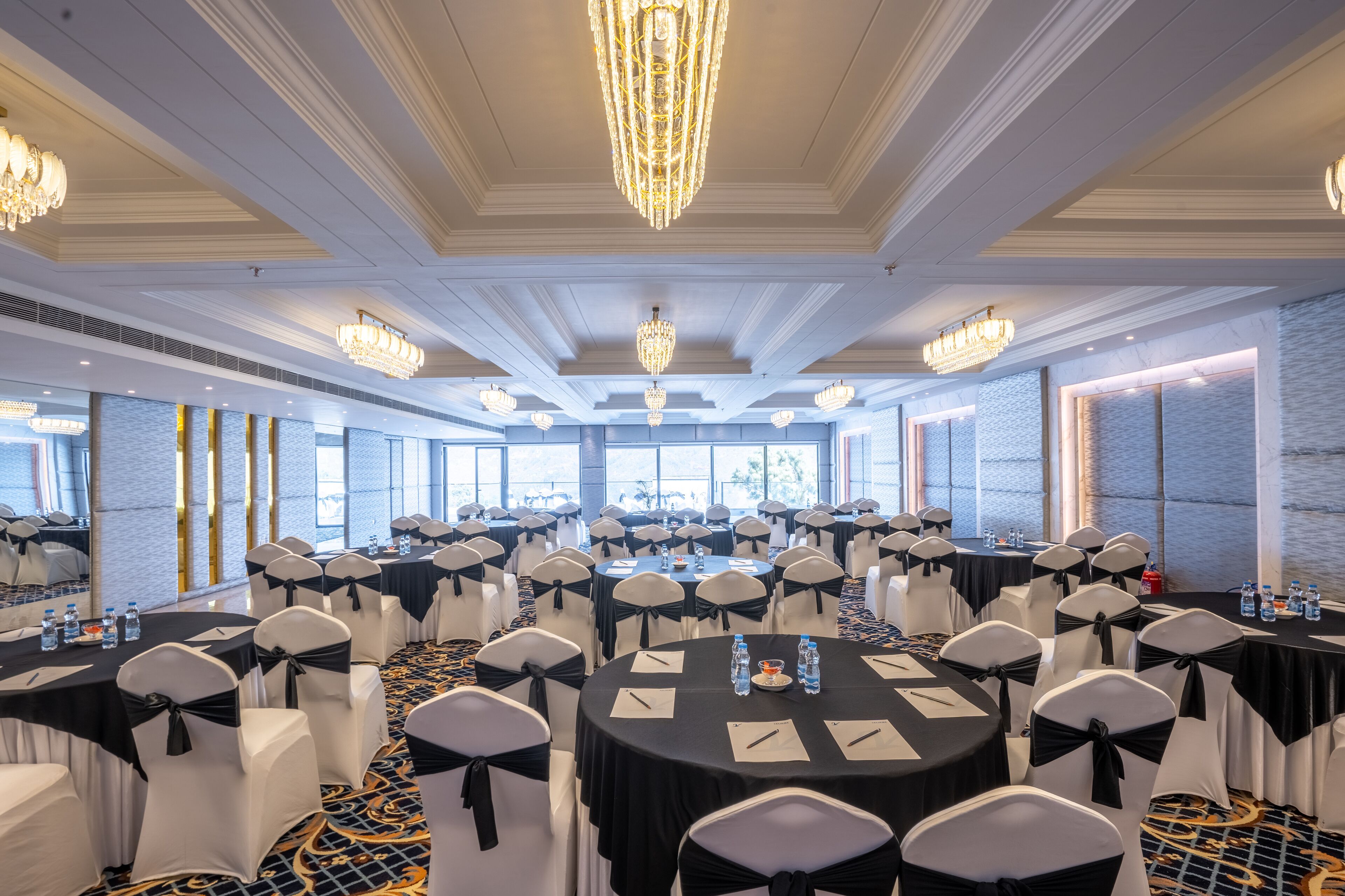 A conference room with round tables, black tablecloths, white chairs with black sashes, chandeliers, and large windows.