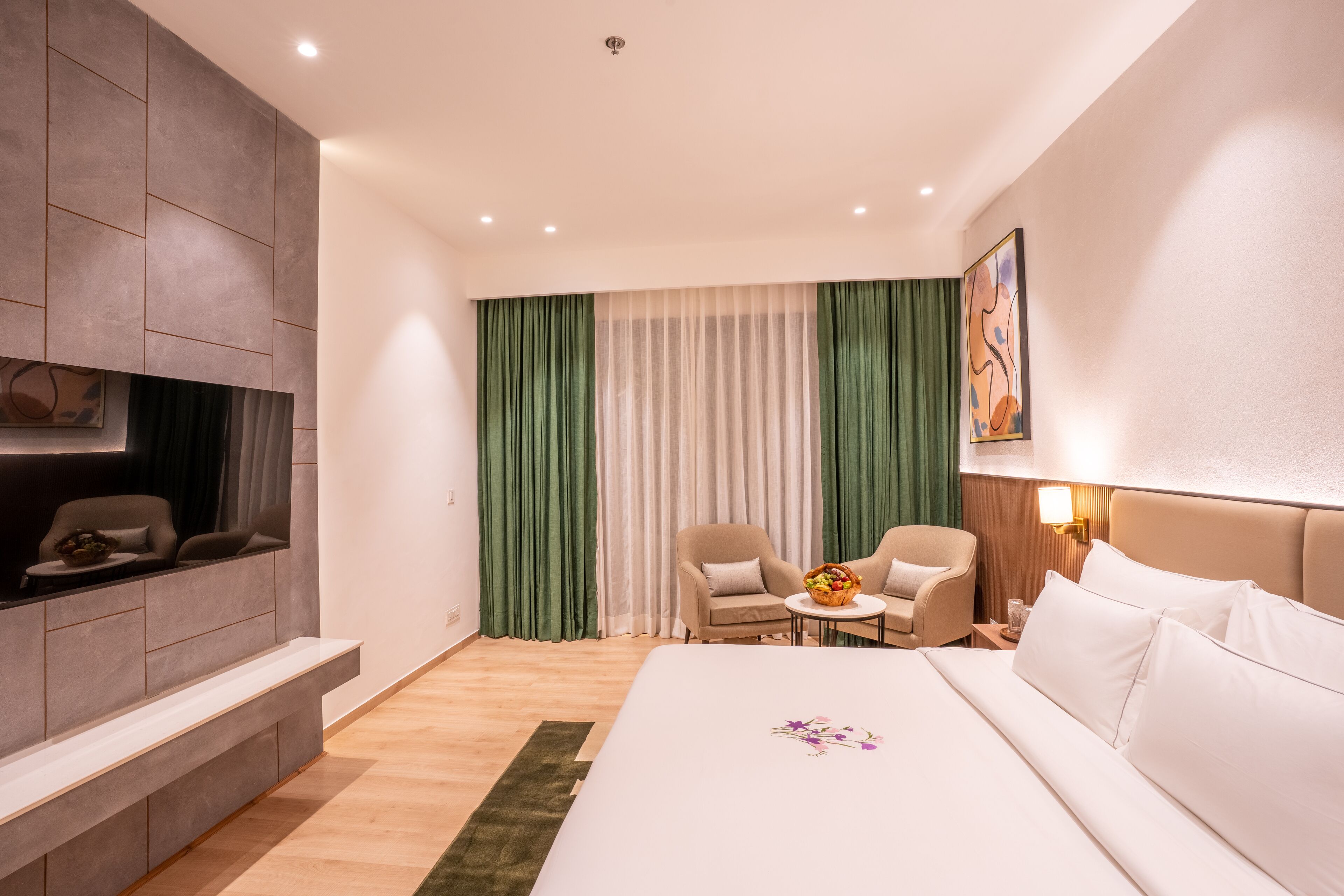 Habitación de hotel con cama, dos sillones, una mesita con una cesta, sofá, televisor montado en la pared y un cuadro en la pared.