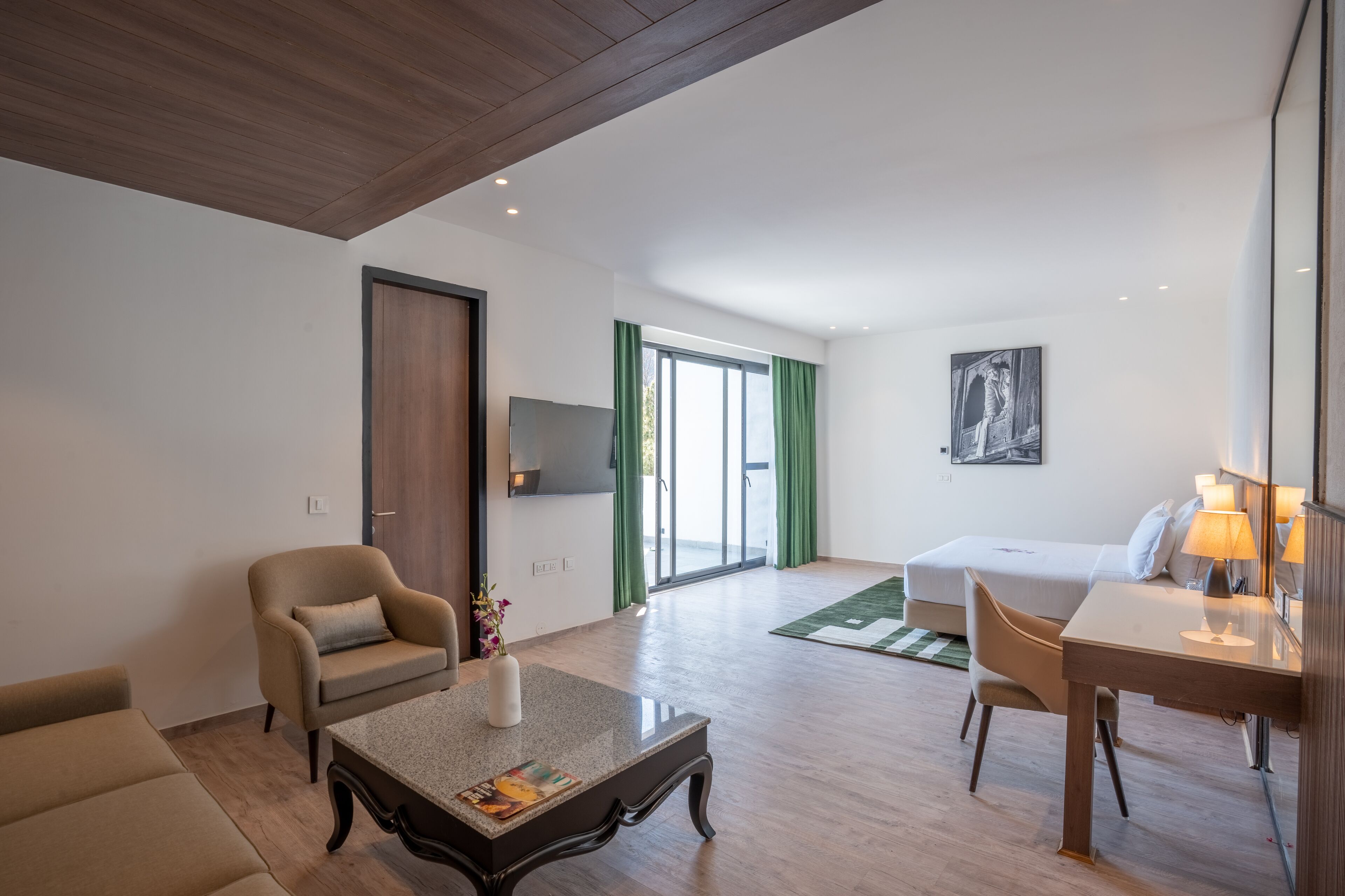 Une chambre d’hôtel moderne avec un plafond en bois, un coin repas comprenant une table et des chaises, un lit agrémenté d’un tapis vert, et un téléviseur fixé au mur.