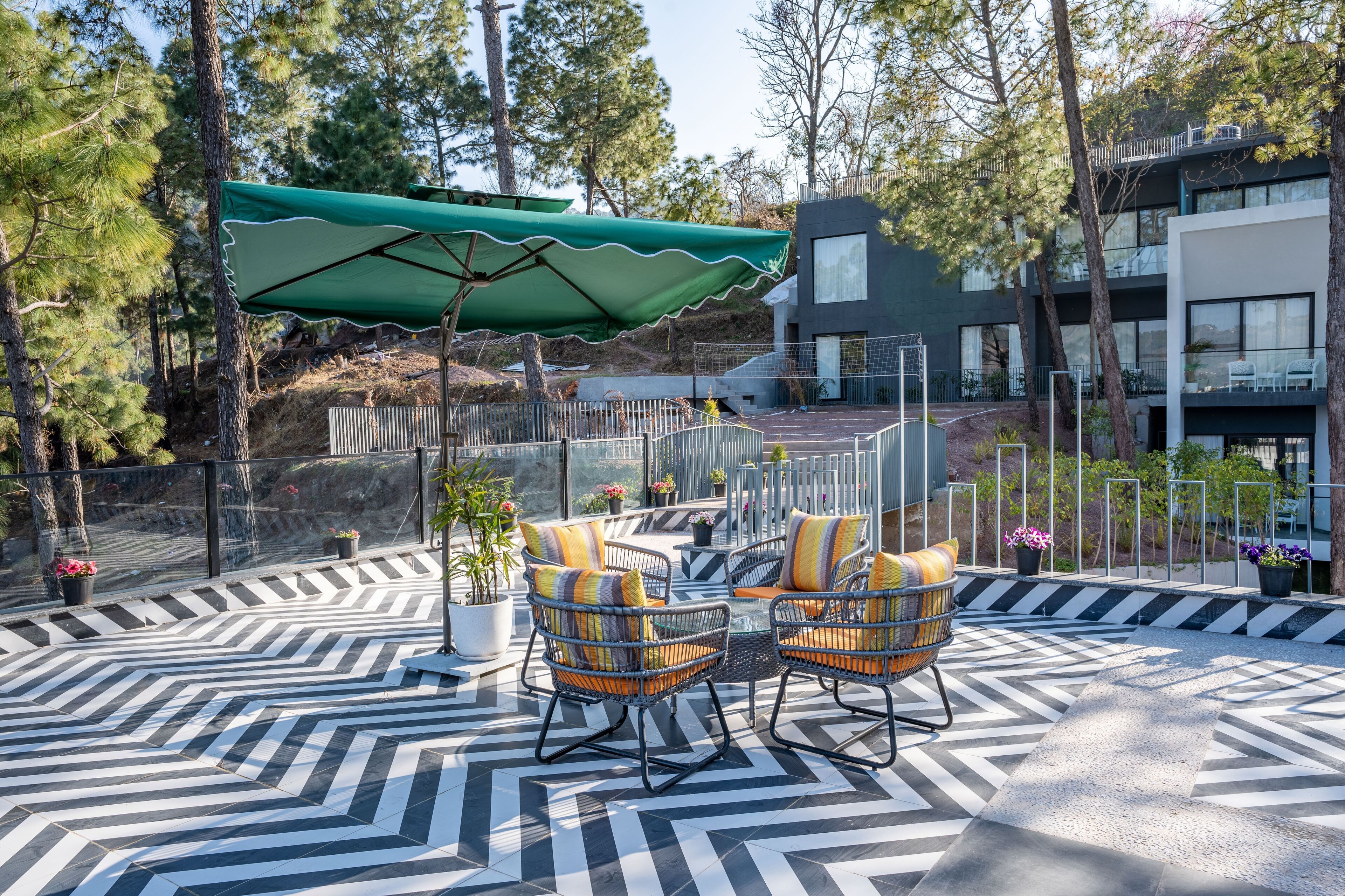 Eine Terrasse im Freien mit einem geometrisch gemusterten Boden, zwei Korbstühlen mit Kissen, einem grünen Sonnenschirm und einem modernen Gebäude im Hintergrund.