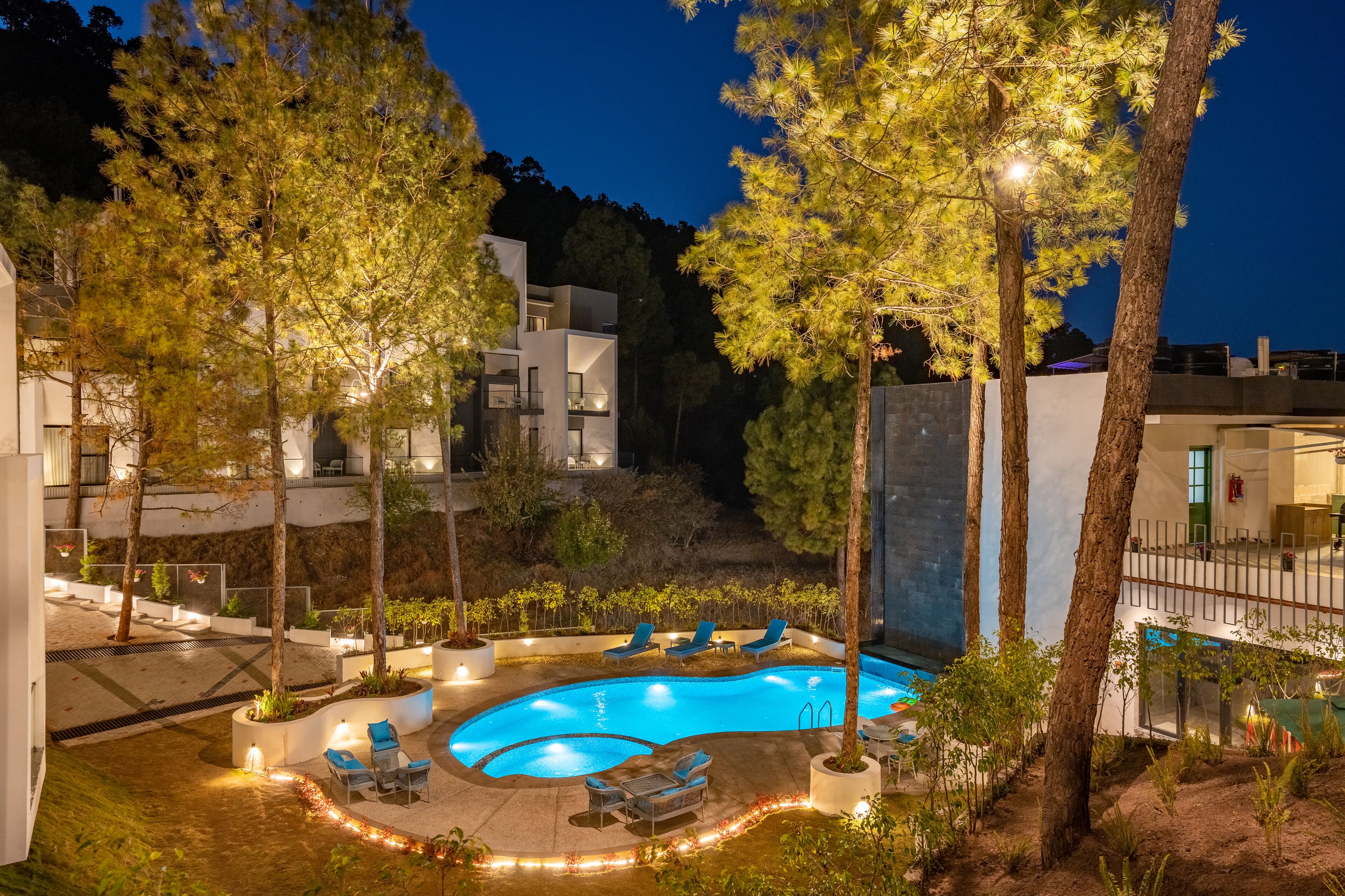 Une piscine entourée d’arbres et de bâtiments modernes, avec des chaises longues à proximité.