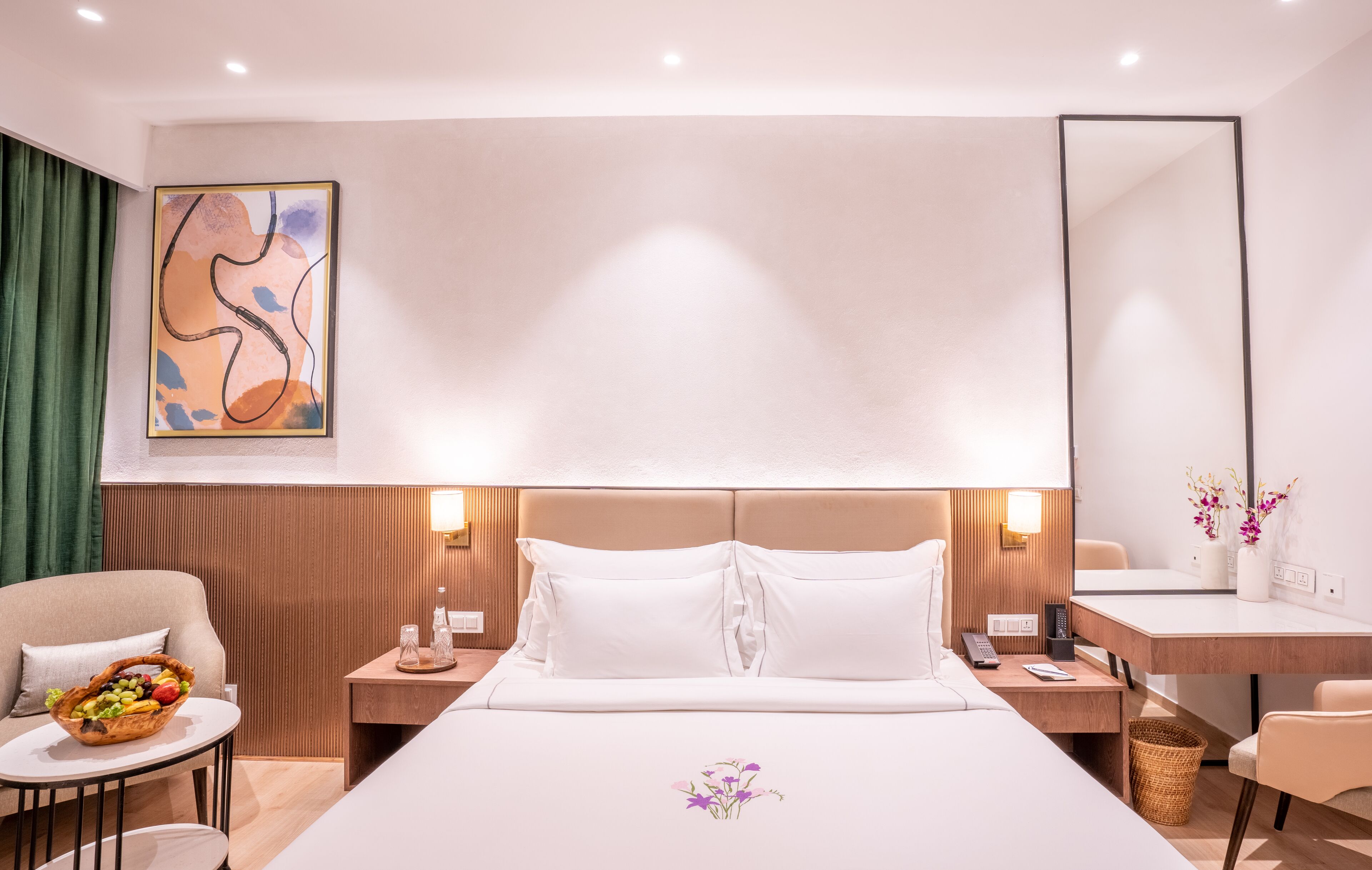 A modern hotel room with a large bed, a bedside table with a lamp, a chair, a small round table with a fruit basket, a desk with a vase of flowers, and a large abstract painting on the wall.