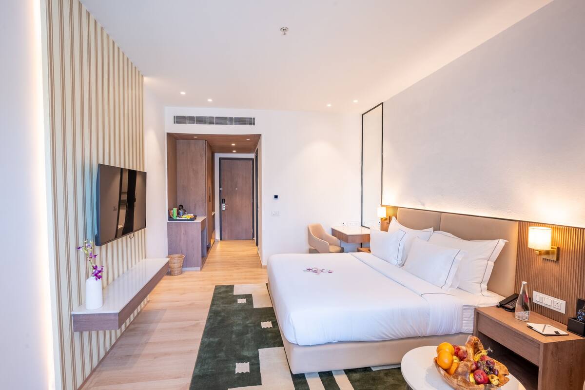 A modern hotel room with a large bed, bedside table, TV, and a small seating area.