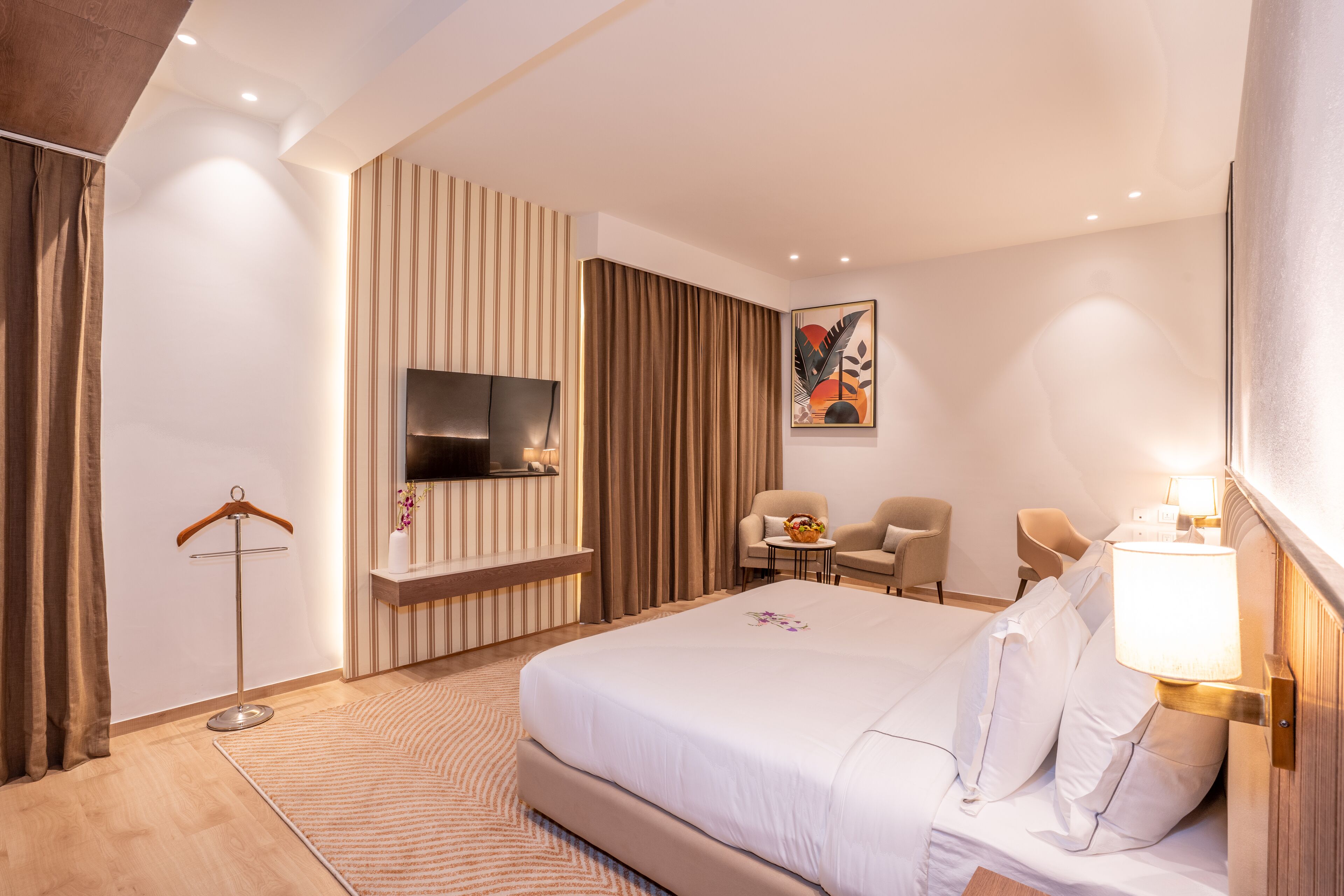 Ein Hotelzimmer mit einem großen Bett, einem flachbildfernseher an der Wand, einer Sitzecke mit zwei Sesseln, einem kleinen Tisch und einer Lampe.