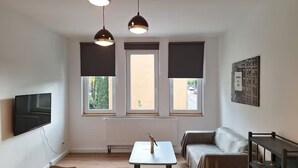 Un moderno salón con un sofá, una mesa de centro y un televisor montado en la pared. Hay tres ventanas con persianas oscuras, un radiador y una lámpara en el techo.