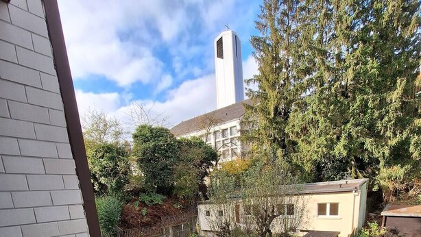 Ein Balkon mit Blick auf einen Garten, eine kleine Gartenhütte und ein hohes Gebäude im Hintergrund.