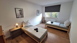 Una habitación con dos camas individuales, una mesita de noche con lámpara, una silla y una ventana con vistas a árboles.