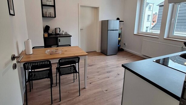 Una cocina compacta con una mesa de madera, sillas negras, un refrigerador y una ventana con vistas a edificios.