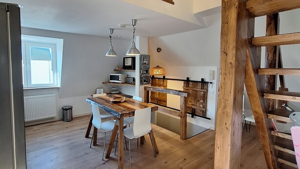 Eine geräumige Küche mit Holzbalken, ein Essbereich mit Holztisch und -stühlen sowie ein Blick auf den oberen Stock.