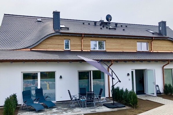 Kaksikerroksinen talo, jossa on kalteva katto, patio ulkokalusteineen ja satelliittiantenni katolla.