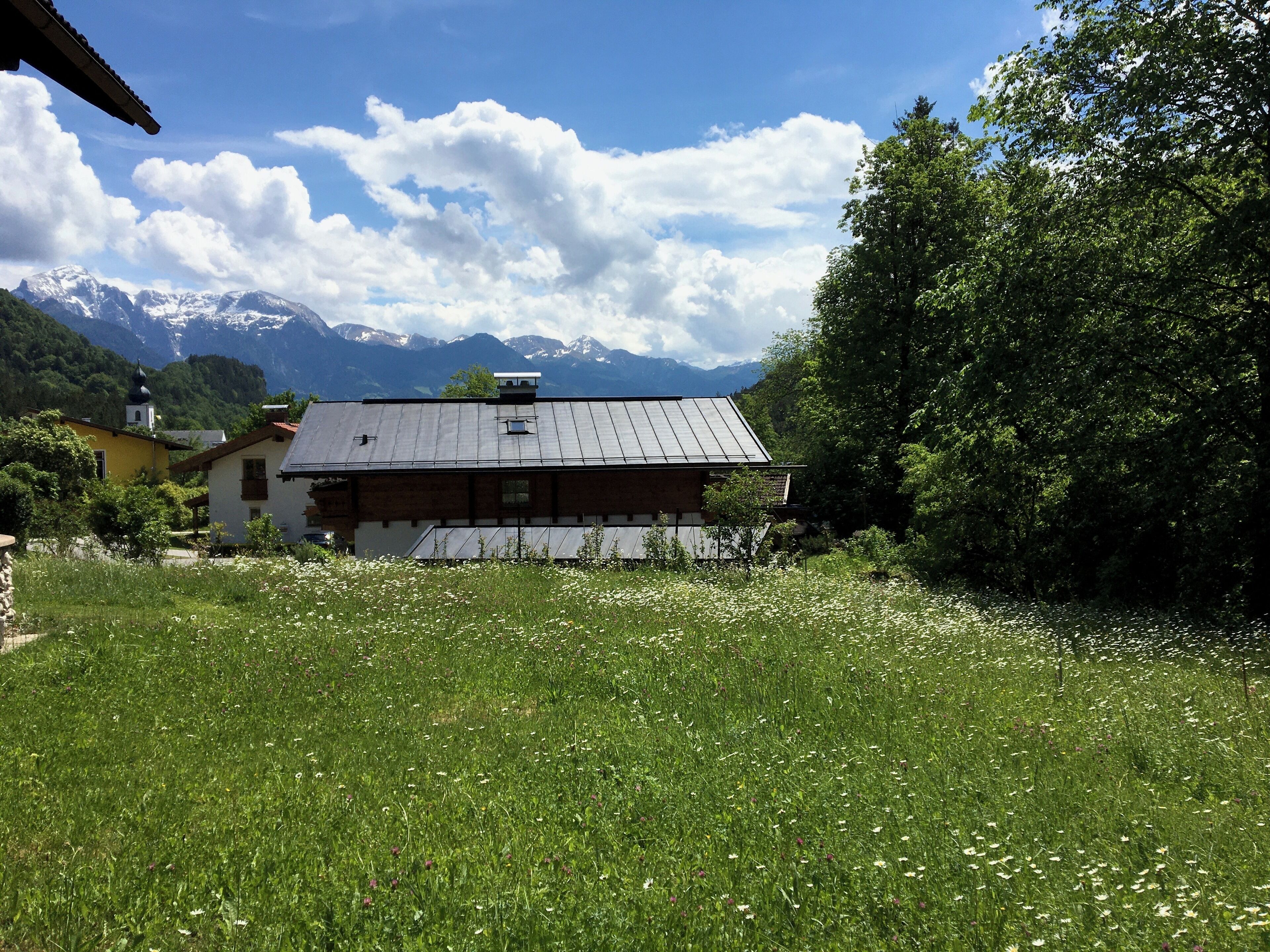 Ein rustikales Gebäude mit geneigtem Dach, umgeben von einer üppigen grünen Wiese und einem Hintergrund aus schneebedeckten Bergen.