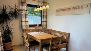 Een eetruimte met een houten tafel en stoelen, een bloempot, een raam met gordijnen en een schilderij aan de muur.