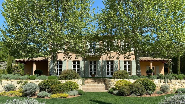 Uma casa de dois andares com fachada de pedra, persianas verdes e um jardim bem cuidado com arbustos variados e gramado.