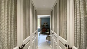 Um corredor com cortinas estampadas, uma cadeira de madeira, um aparador e um quadro na parede.