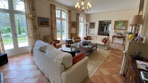 Uma sala de estar ampla com um sofá grande, uma mesa de centro e um candelabro. Há várias janelas com cortinas, um quadro na parede e uma porta que dá para o exterior.