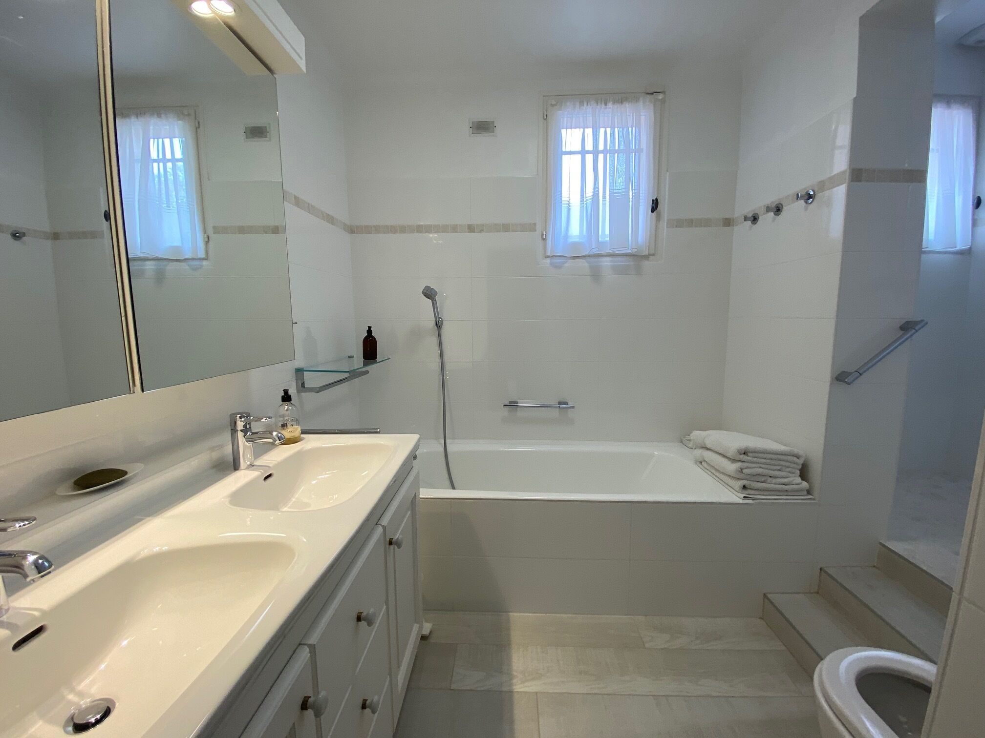 A bathroom with a double sink, a bathtub, and a toilet. There is a mirror above the sink and a window with curtains.
