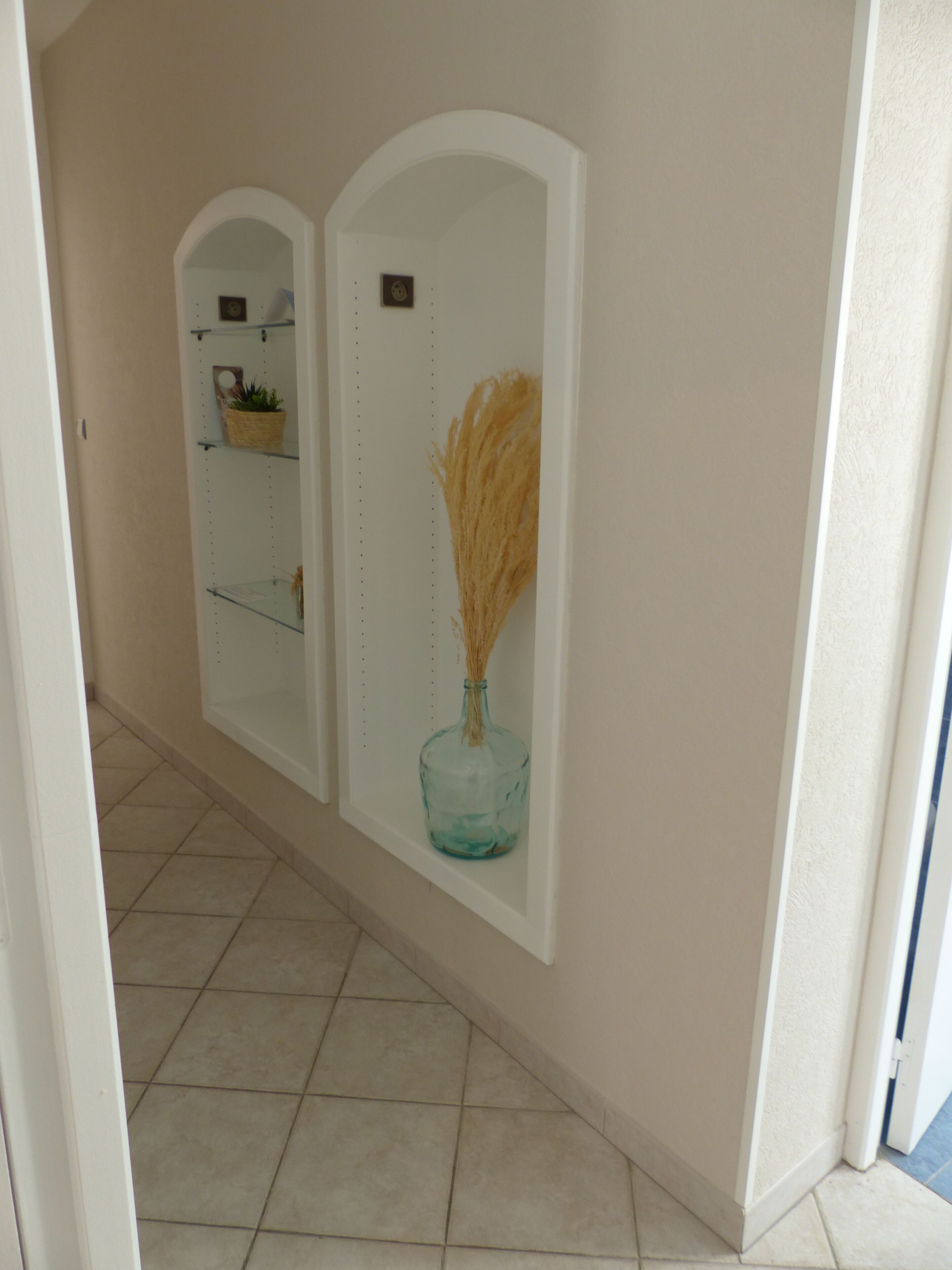A hallway with a tiled floor, a white wall with a decorative mirror, and a blue vase with dried plants.