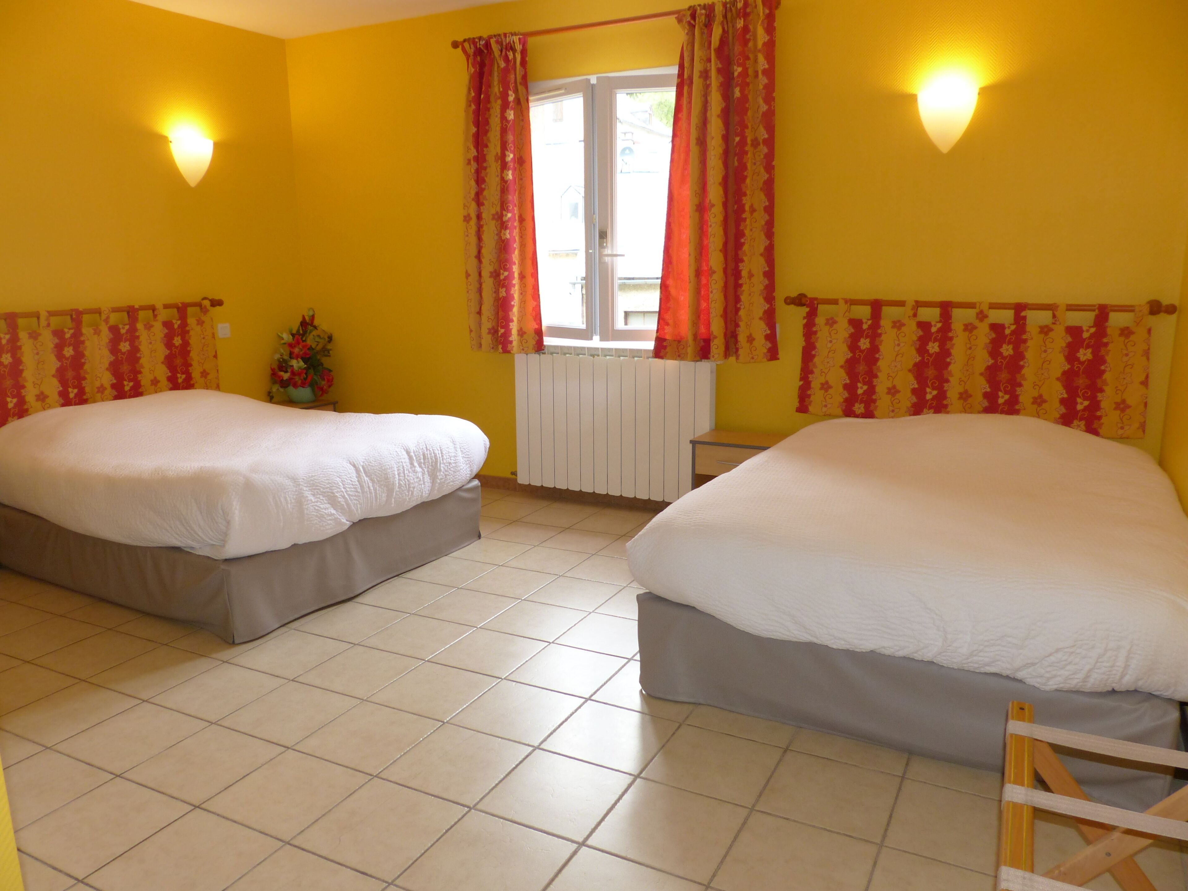 A room with two beds, a window with curtains, a radiator, and a small table with a plant.