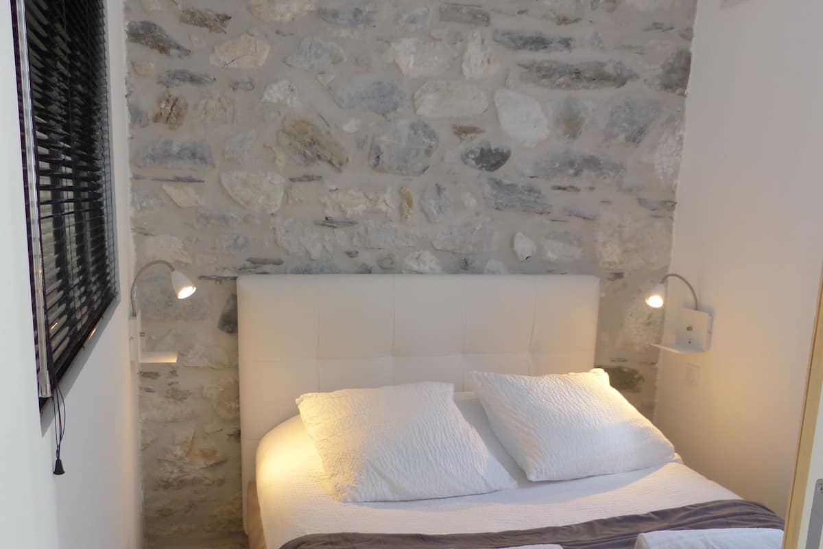 A bedroom with a stone wall, a white bed with grey bedding, two bedside lamps, and a wooden floor.