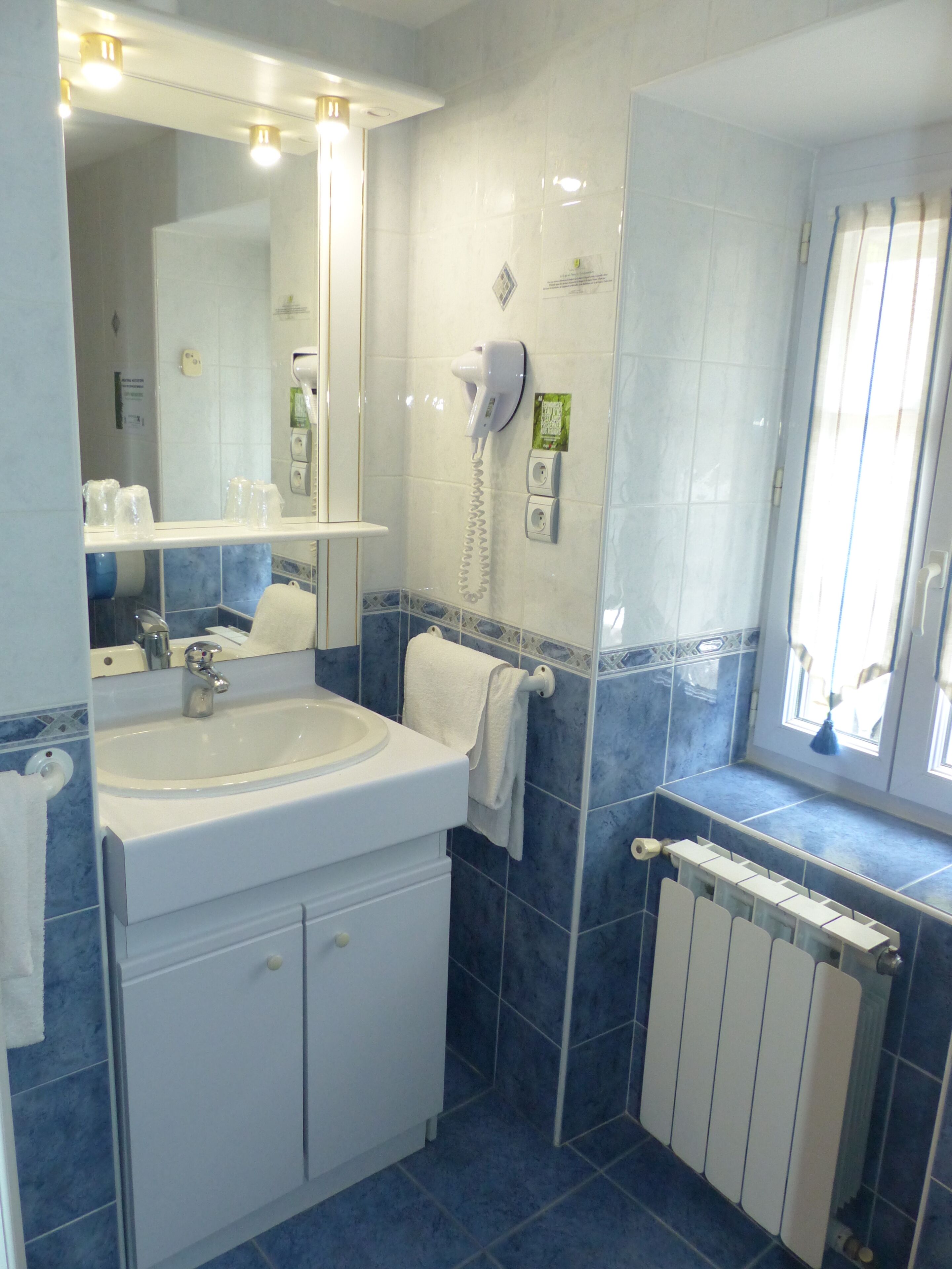 Casa de banho com lavatório branco, espelho, porta-toalhas, secador de cabelo e janela com persianas.