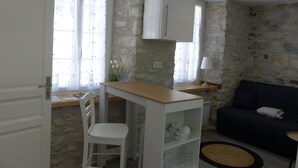A compact living space with a kitchenette, a dining area with a white chair, a sofa, and a stone wall. - Hotel Auberge de l'Isard (Saint-Lary)
