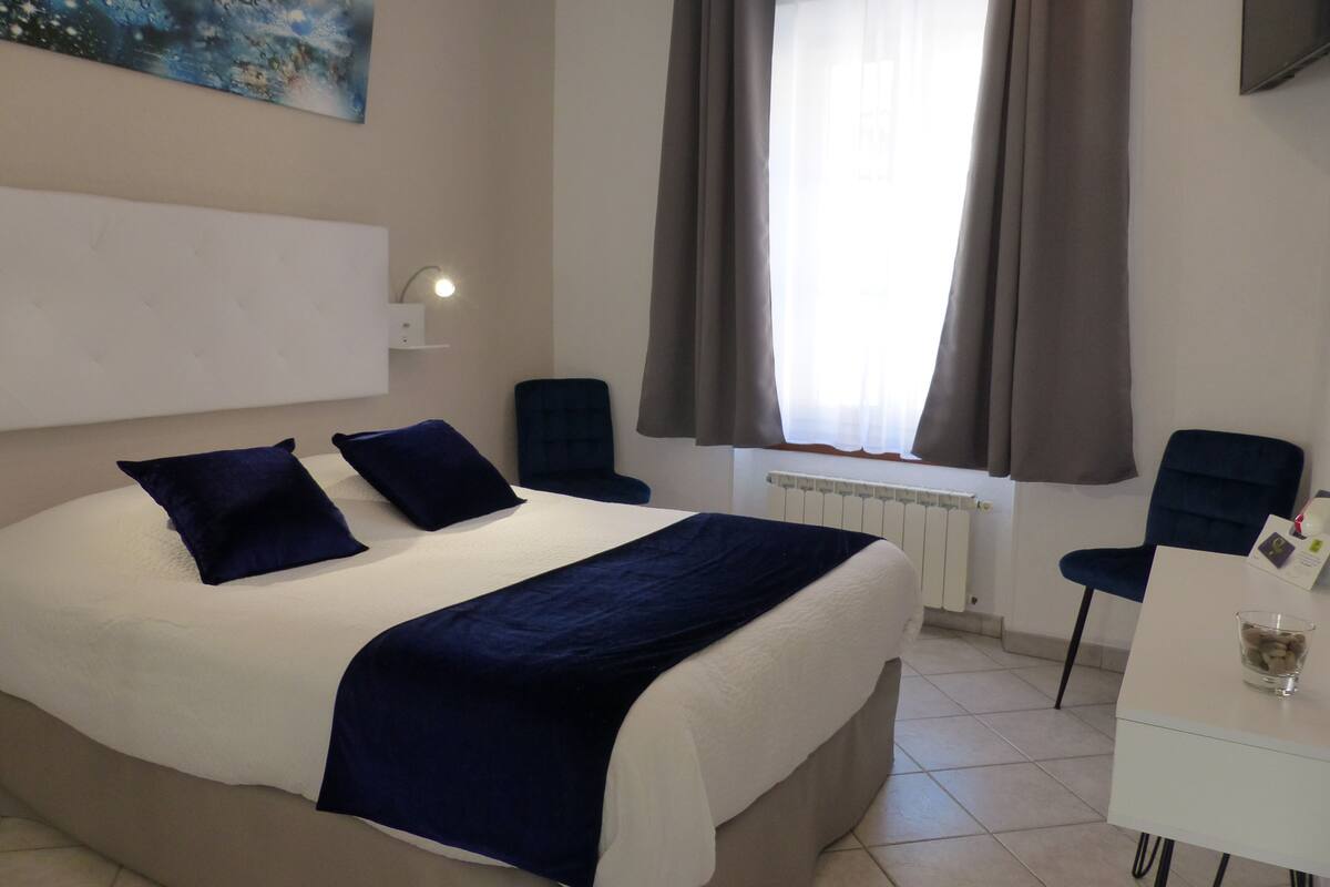 A hotel room with a bed, a desk, a chair, a window with curtains, a radiator, and a painting on the wall.