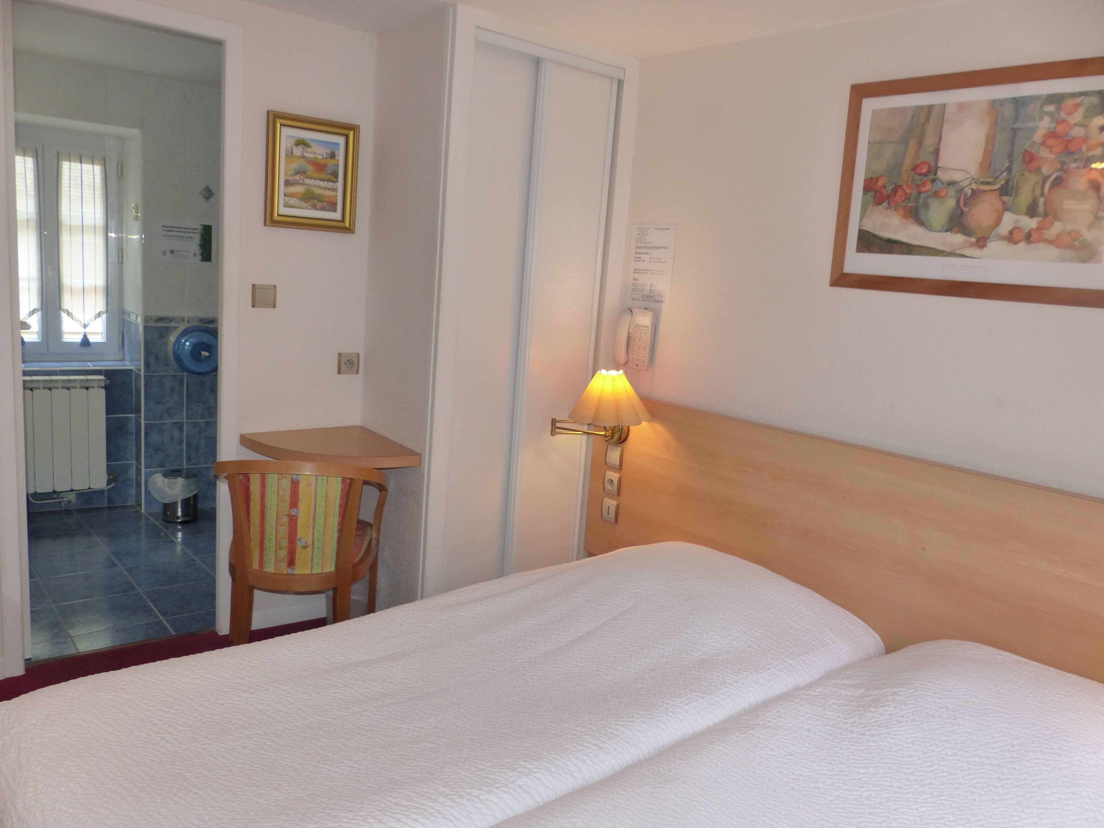 Una habitación de hotel con cama, escritorio, silla, lámpara y un cuadro en la pared.