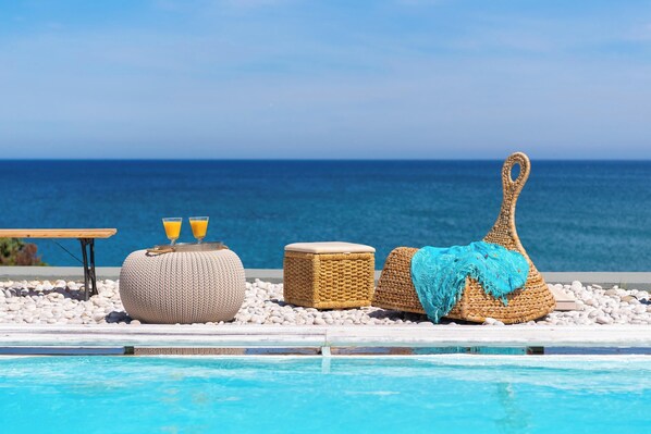 A poolside area with wicker furniture, a table with drinks, and a view of the sea.