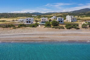 A coastal area with modern white buildings, clear turquoise waters, and a hilly landscape in the background.