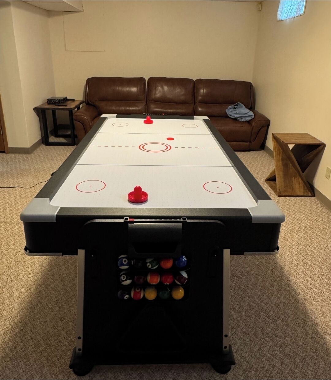 Une table de jeu de shuffleboard dans une pièce où se trouvent un canapé en cuir marron et une petite table d’appoint en bois.
