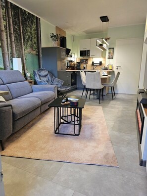 Un salon moderne avec un canapé gris, une table basse et un coin repas comprenant des chaises, et une cuisine en arrière-plan.