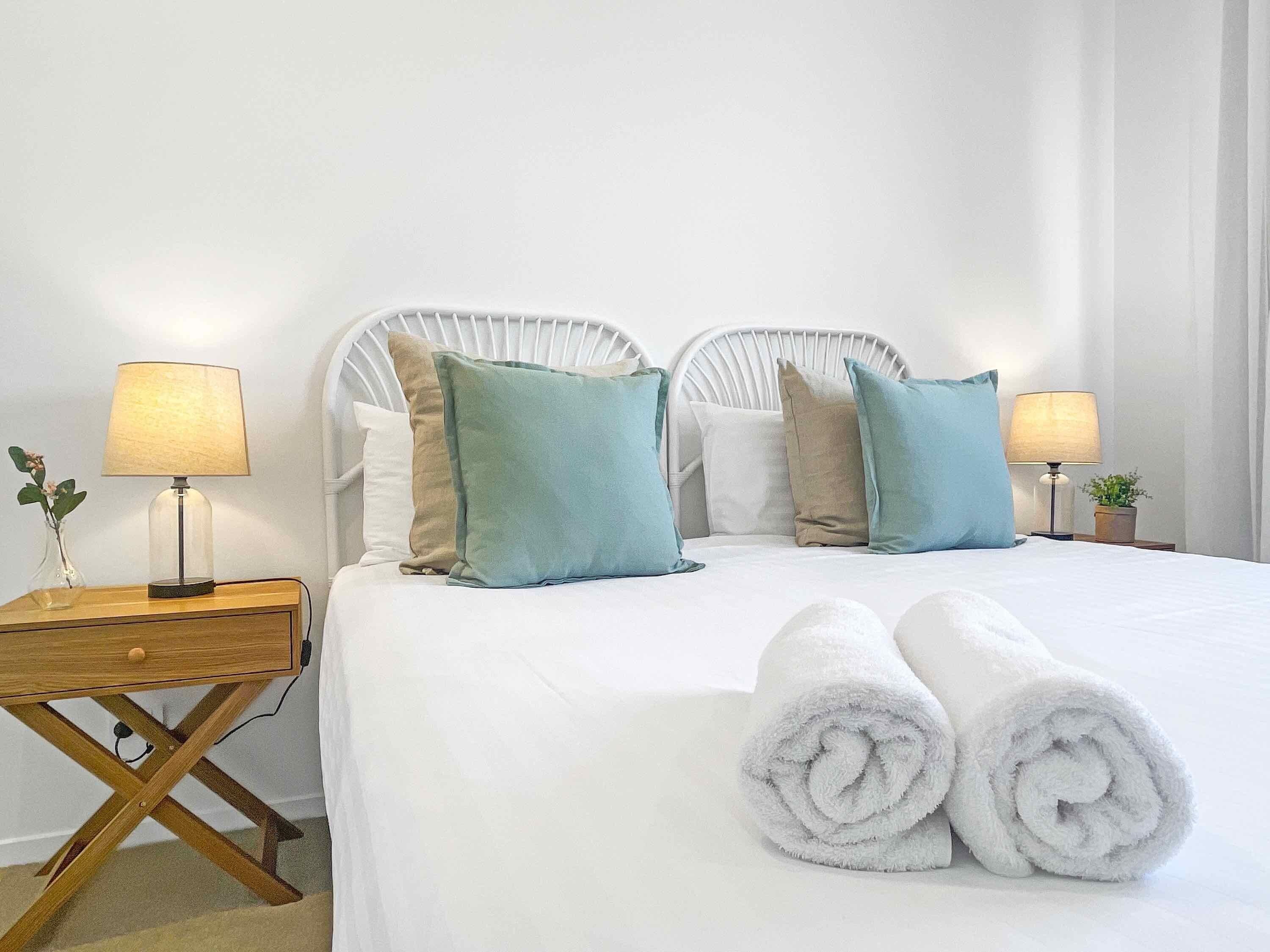 Cama bem arrumada com roupa de cama branca e dois travesseiros verde-água, mesa de cabeceira de madeira com abajur, e uma toalha branca enrolada sobre a cama.