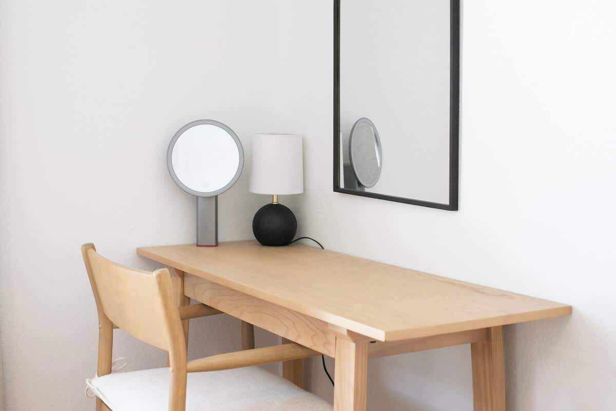 Ein Holztisch mit einem Stuhl, einem runden Spiegel und einer Lampe an einer weißen Wand.