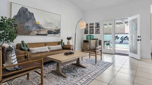 A modern living room with a sofa, coffee table, and chairs, a large painting on the wall, and a view through a glass door.
