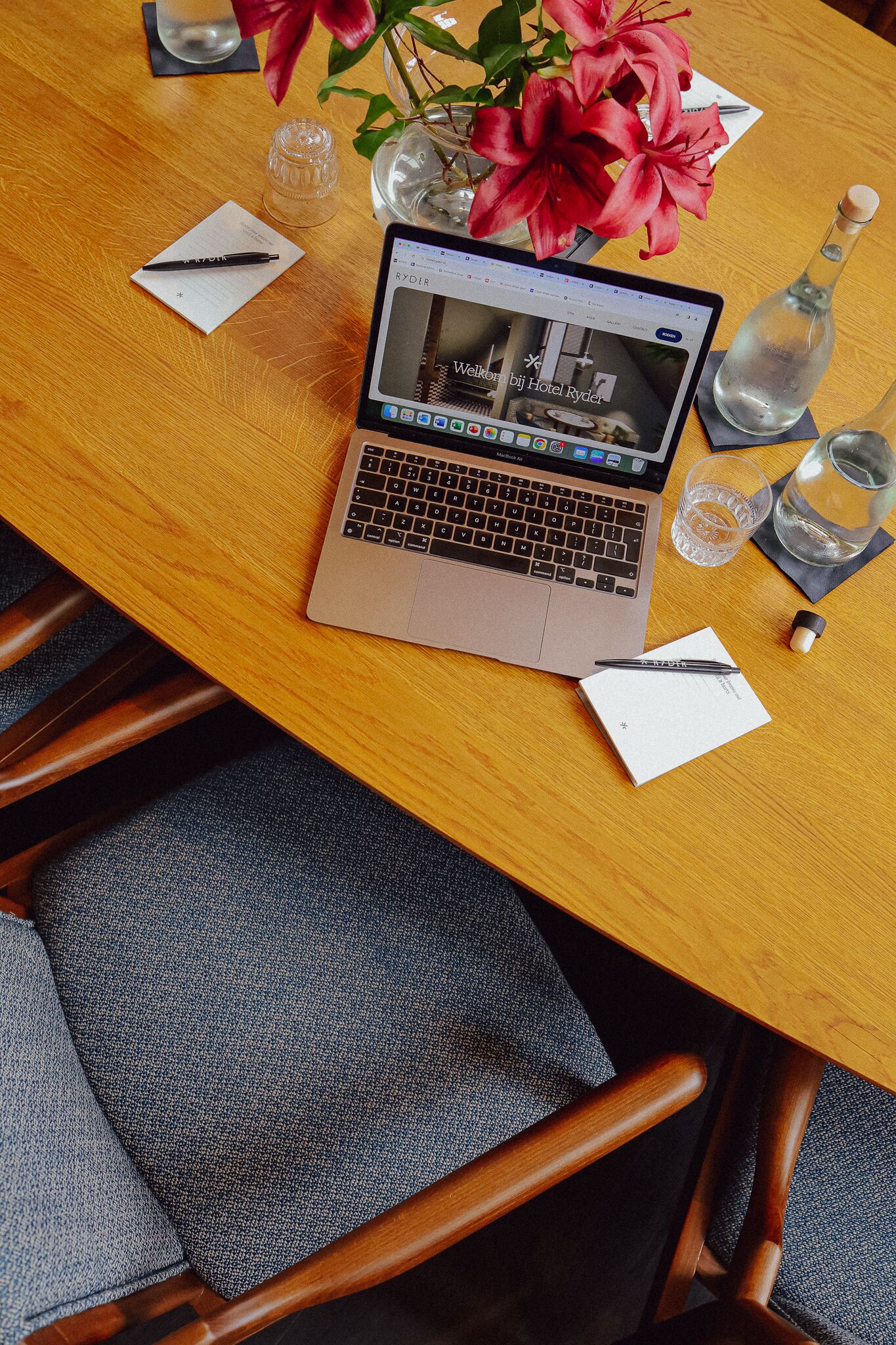 A laptop on a wooden table with a vase of red flowers, a glass of water, and a bottle.