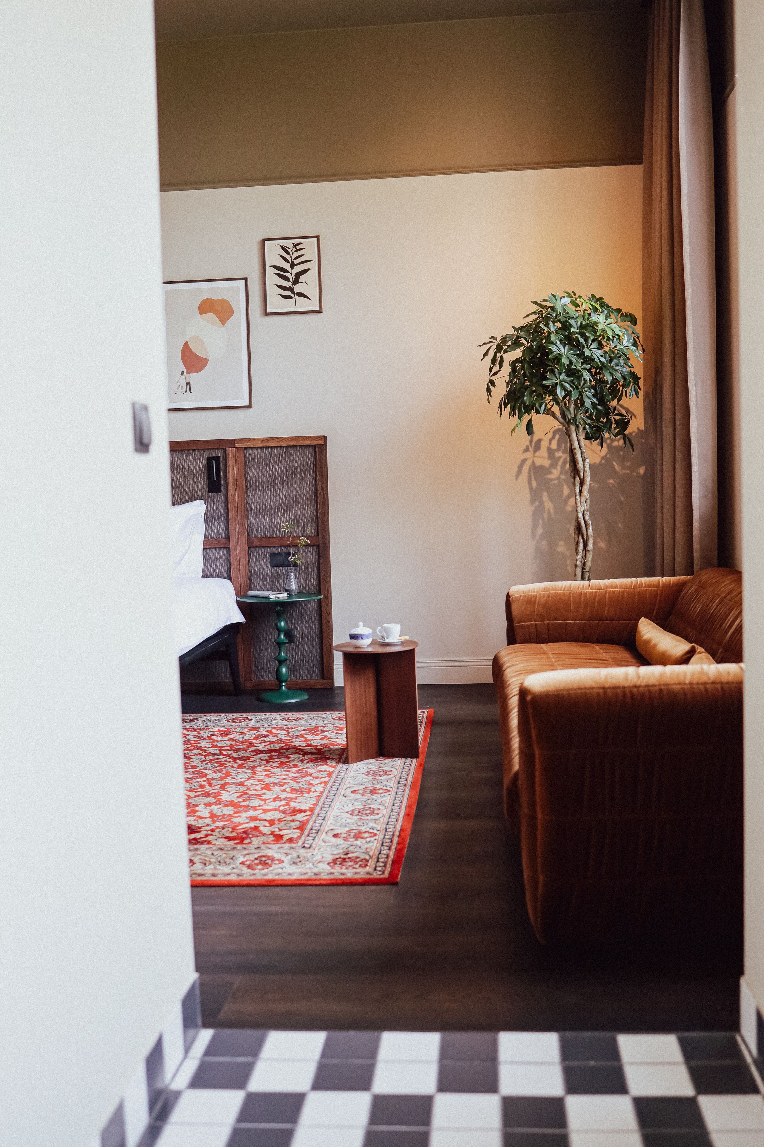 A hotel room with a bed, a green side table, a brown leather sofa, a red and white patterned rug, and a tall plant.