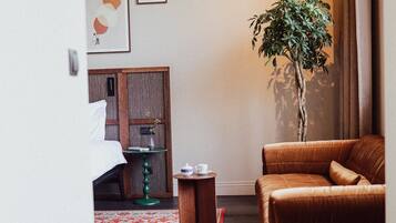 A hotel room with a bed, a green side table, a brown leather sofa, a red and white patterned rug, and a tall plant.