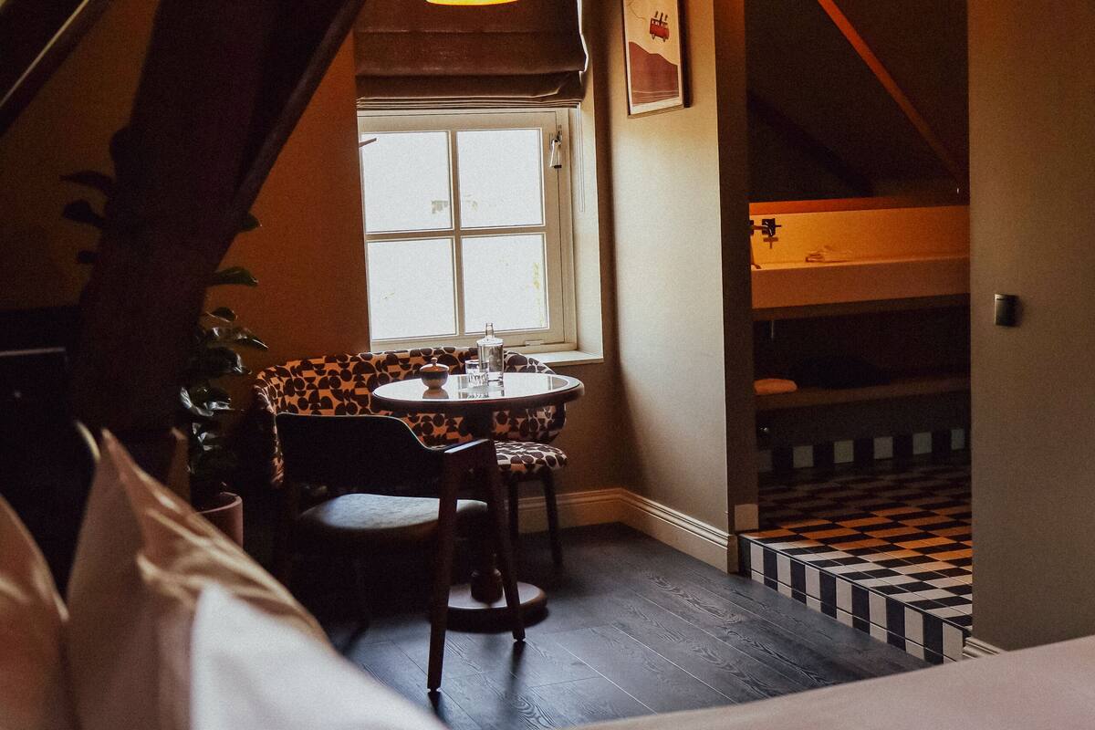 A bedroom with a bed, a window, a table with chairs, a staircase, and a hanging plant.