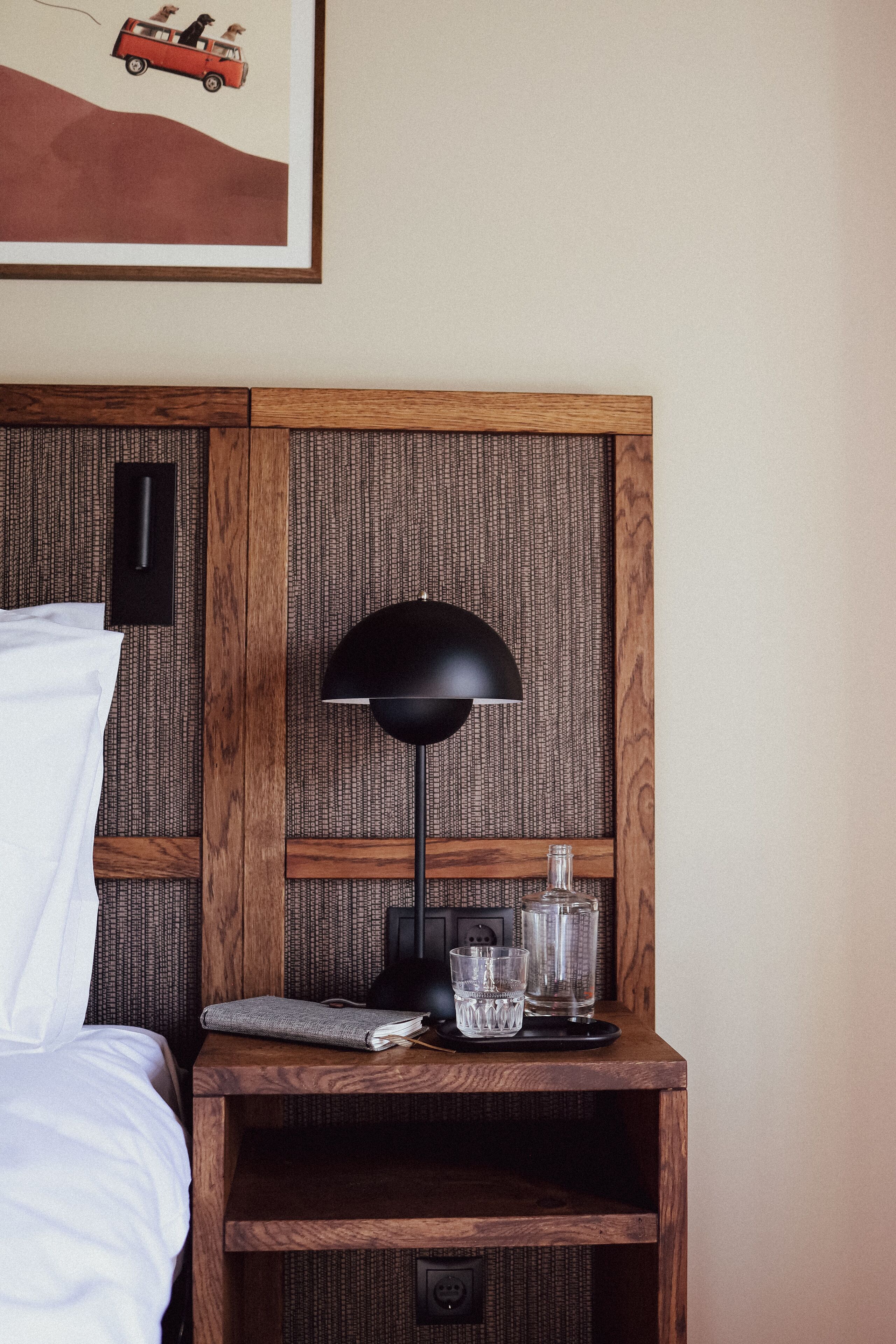 A neatly made bed with white linens, a wooden nightstand with a black lamp, glasses, and a book, and a framed picture above the nightstand.