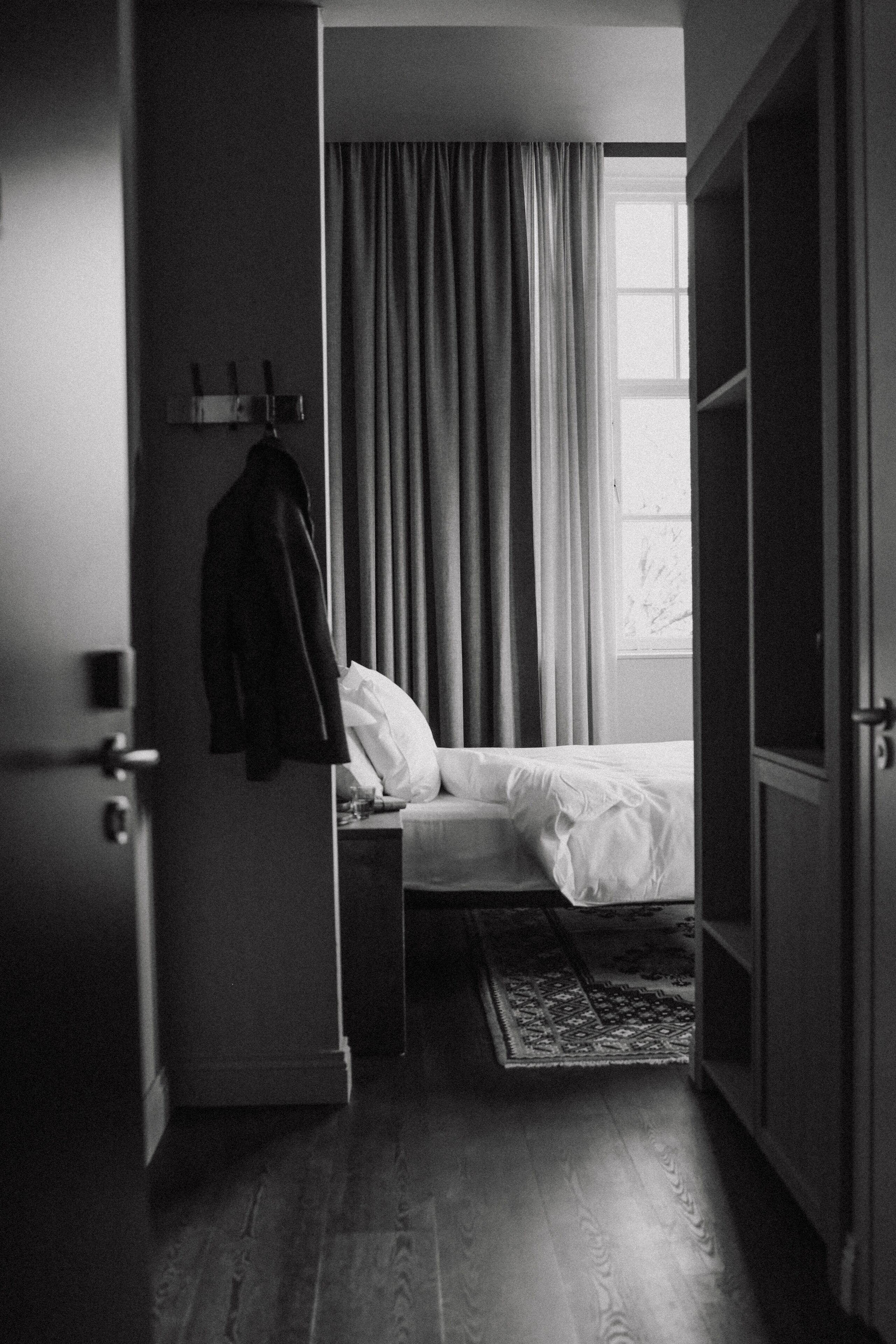 Een hotelkamer met een bed, nachtkastje, hangende jas, raam met gordijnen en een houten vloer.