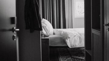 Een hotelkamer met een bed, nachtkastje, hangende jas, raam met gordijnen en een houten vloer.