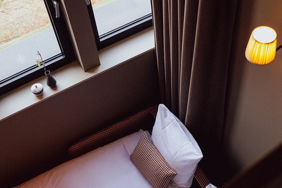 A small, cozy room with a bed, a window with curtains, a lamp, and a view of the outside.