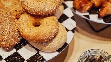 Una mesa con diferentes productos horneados, como bagels y croissants, un frasco de crema de chocolate y un mantel a cuadros.