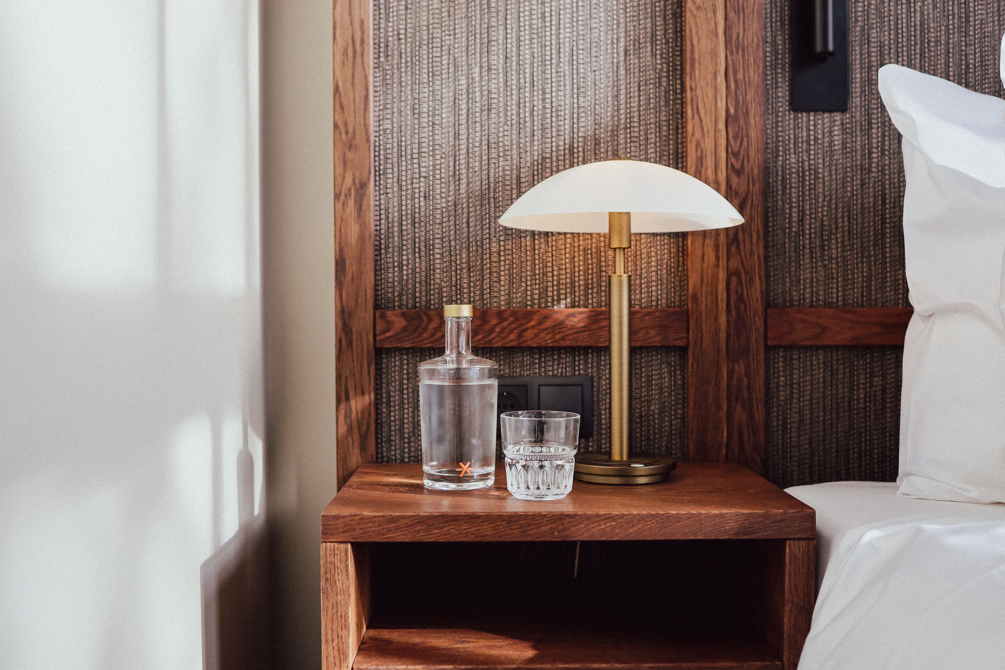 A wooden bedside table with a clear bottle, a glass, and a lamp.