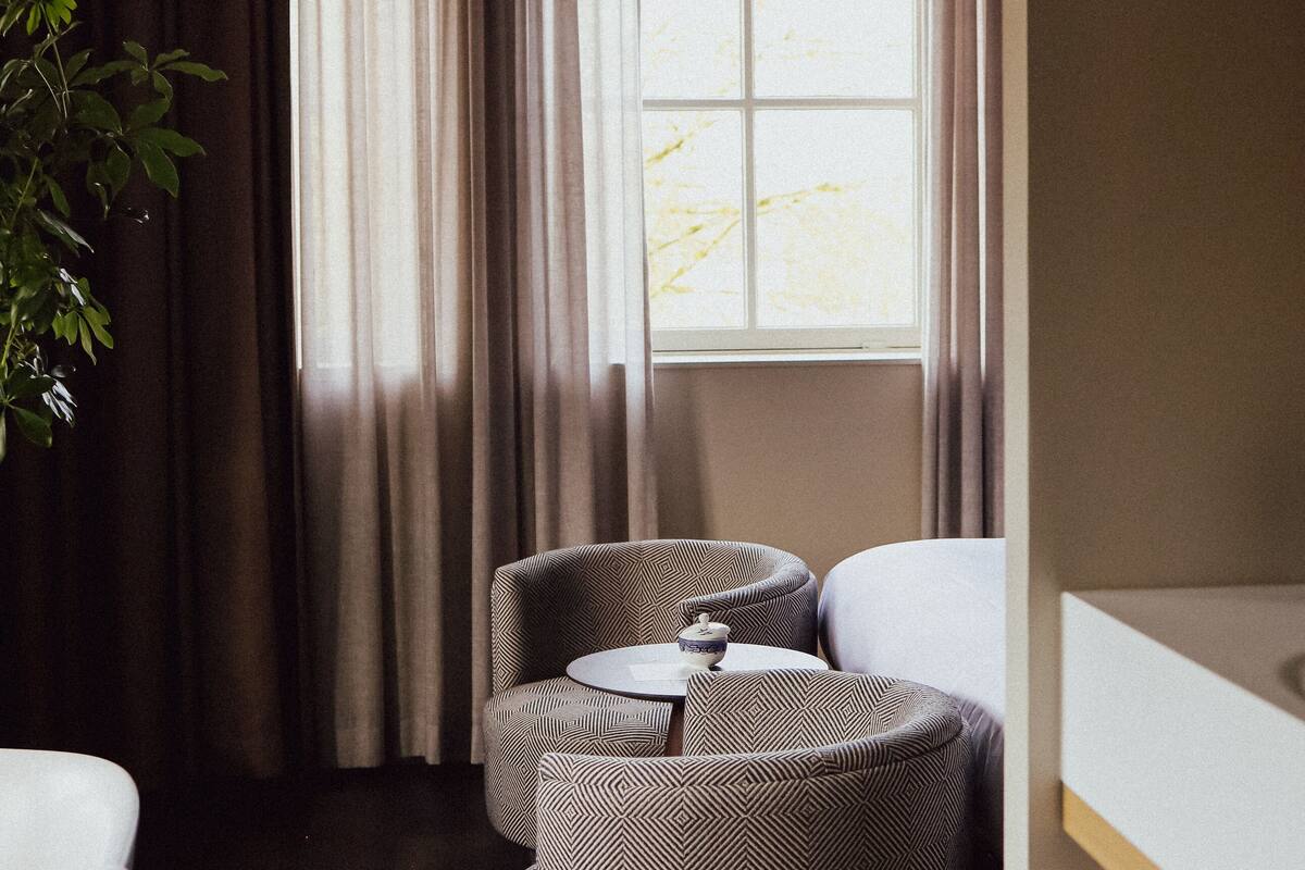 A hotel room with a dark wooden floor, a large window with curtains, a sofa, a small table with a cup, and a bedside table with towels.