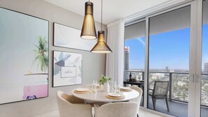 A modern dining area with a round table set for four, two pendant lights, and a view of the city through large windows. - Exquisite 1BR High-Rise | Sunset Views (Hollywood)