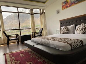 A bedroom with a large bed, two chairs, a view of mountains, and a window with curtains. - Hotel The White Castle (Kargil)