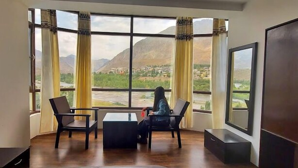 A room with a large window offering a view of a mountainous landscape and a river. There are two chairs and a small table in the room. - Hotel The White Castle (Kargil)
