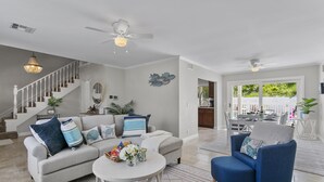 A spacious living room with a grey sofa, blue armchair, and a white coffee table. A staircase with a white railing leads to another level. - Stunning Coastal Escape w. private pool & spa, arcade, near themed parks & beach (Mission Viejo)