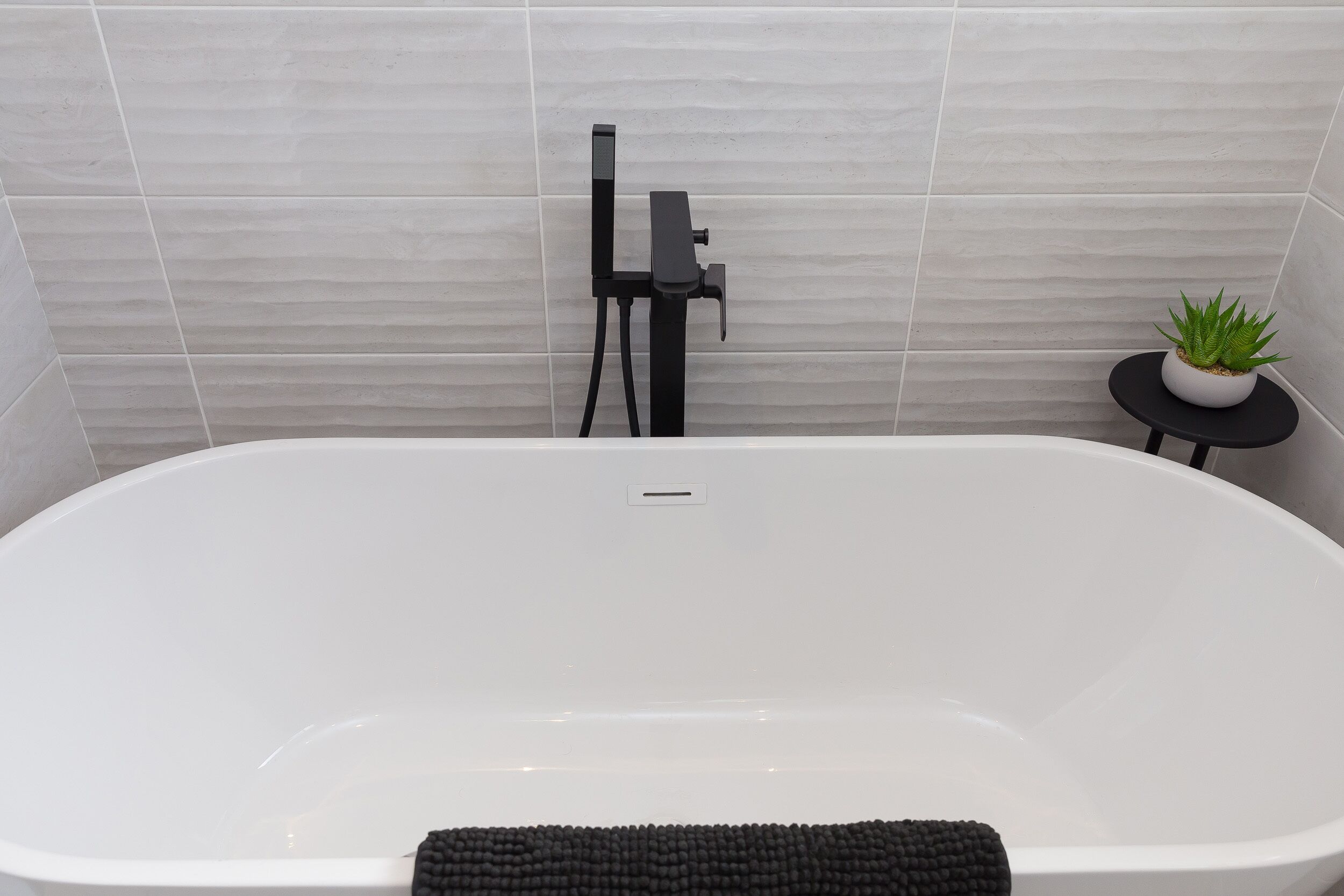 A modern bathroom with a white bathtub, a black faucet, and a small potted plant on a black stool.